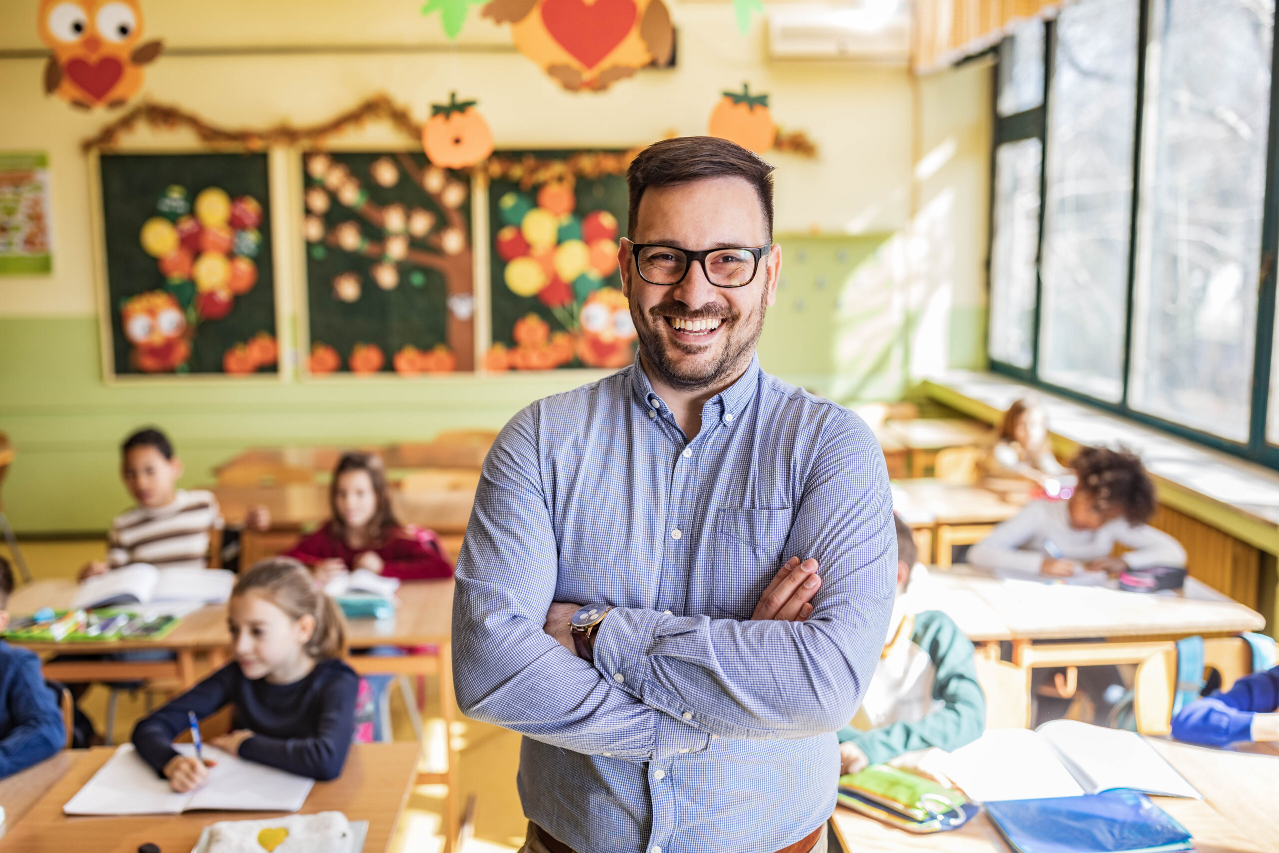 Young happy teacher standing on a class with his arms crossed and looking at camera. Elementary students are in the background.