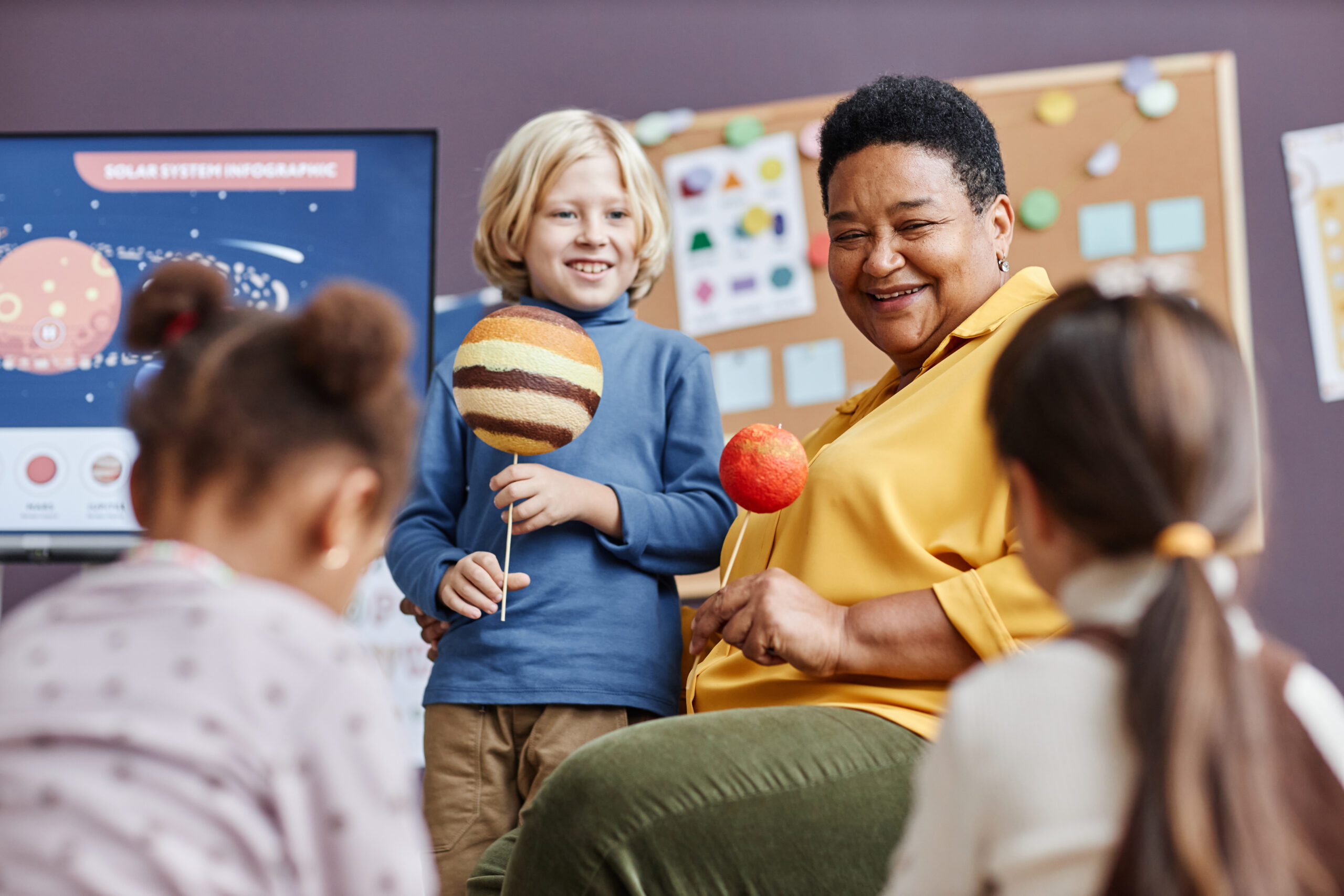 Focus on happy mature teacher and cute little boy with planet models during discussion of solar system at lesson in kindergarten