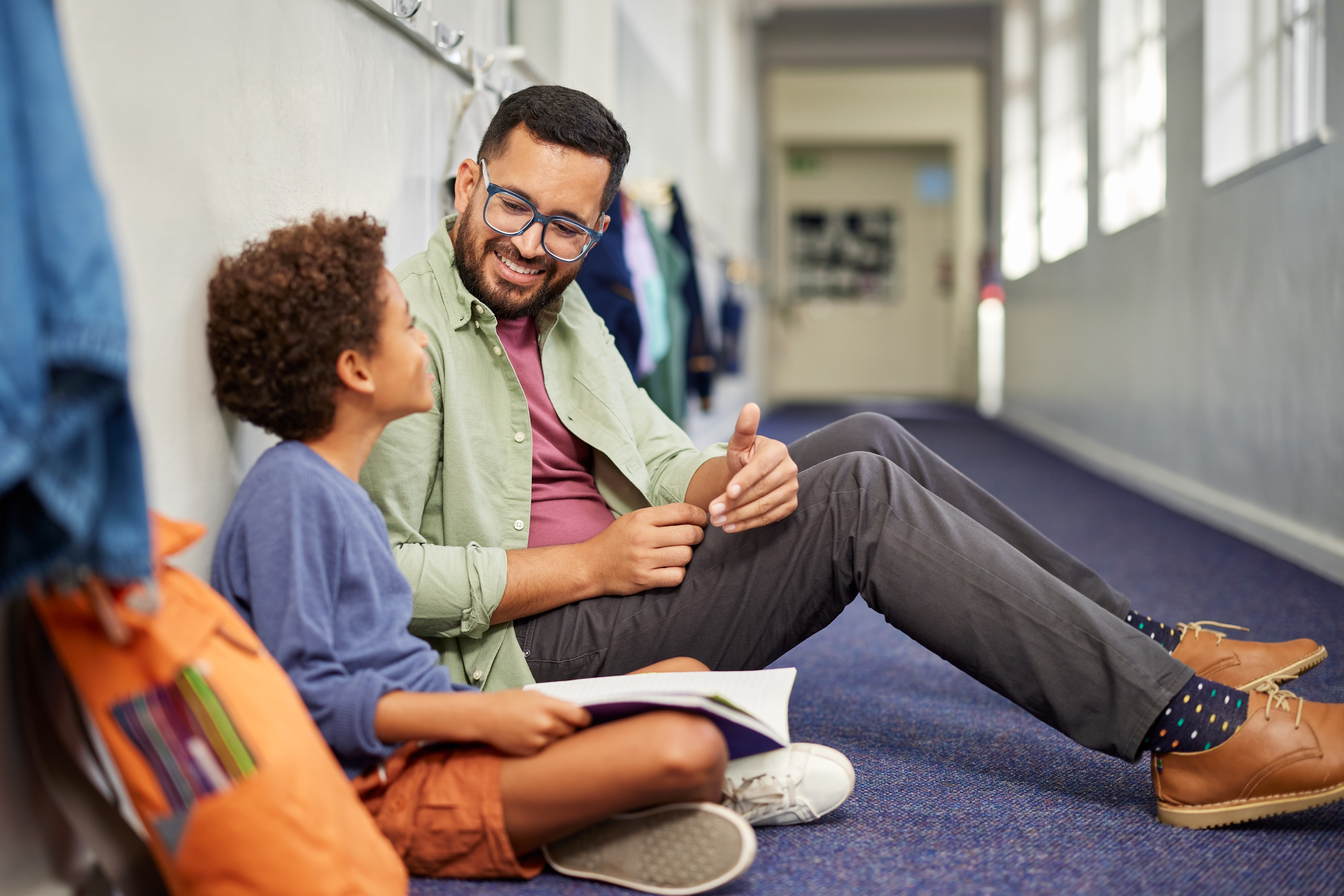 Young male teacher speaking to studying boy in school while sitting on floor in corridor. Man working with a child while discussing about examination doubts. Young and friendly teacher speaking to boy in school hallway while helping him in studies.